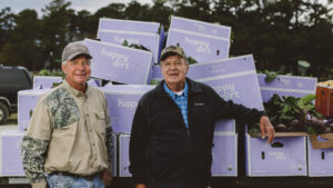Happy Dirt farmer-owners Owen and Vernon Rouse stand in front of a trailer stacked with purple Happy Dirt boxes filled with organic cabbage and beets. Both are smiling, dressed in casual farm clothing, with trees and a truck in the background.