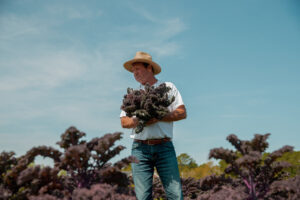 Happy Dirt organic farmer-owner Herbie Cottle standing in a North Carolina kale field, holding freshly harvested bunches of organic kale under a bright blue sky.