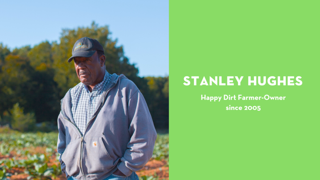 Stanley Hughes, a Happy Dirt farmer-owner, stands in a field on a sunny day wearing a gray hoodie and checkered shirt. Trees and crops stretch behind him under a clear blue sky. Text beside him reads “Stanley Hughes – Happy Dirt Farmer-Owner since 2005” on a bright green background.
