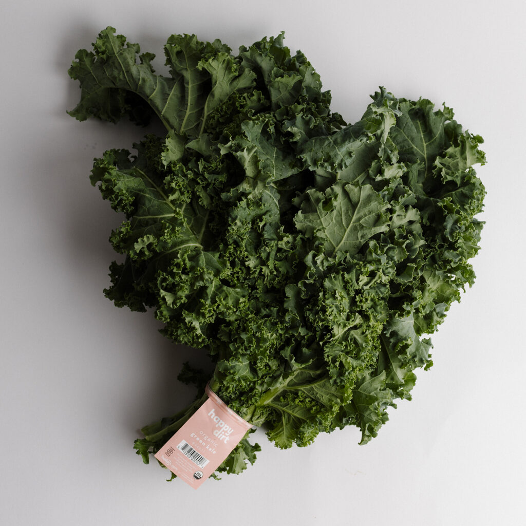 A fresh bunch of Happy Dirt organic green kale with curly, dark green leaves, wrapped in a peach-colored Happy Dirt label featuring the USDA Organic seal, barcode, and website on a clean white background.