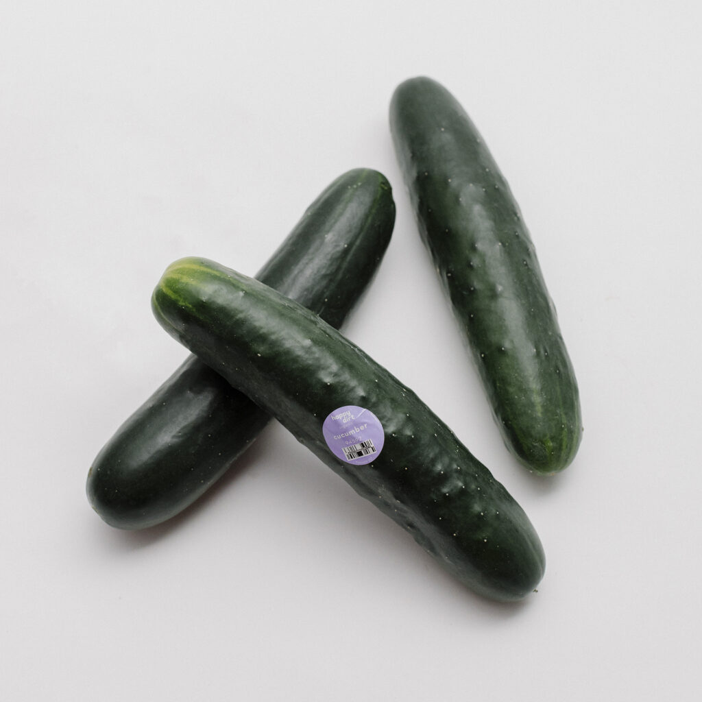 three organic slicing cucumbers, one with a purple happy dirt sticker, laying on a white surface.