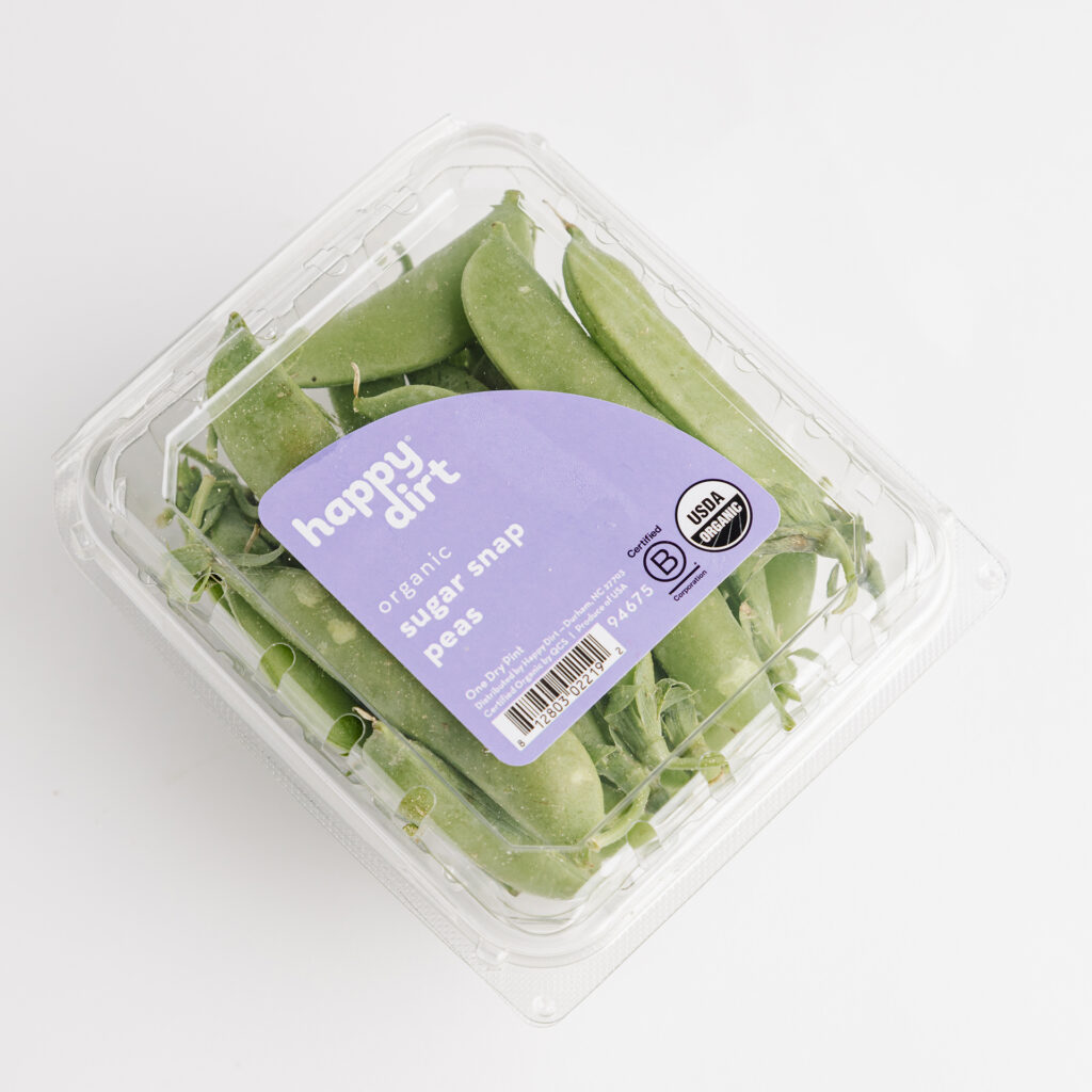 a clamshell pint is filled with organic sugar snap peas with a purple happy dirt sticker on top, laying on a white surface.
