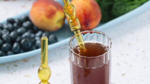 Two glasses of iced sun tea with amber glass straws on colorful coasters, in front of a tray with fresh organic blueberries, peaches, and basil, capturing a bright, summery kitchen scene.
