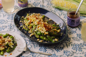 Charred organic shishito peppers with corn salad, Tin Man Turkish chili oil, toasted peanuts, and benne seeds served on a platter.